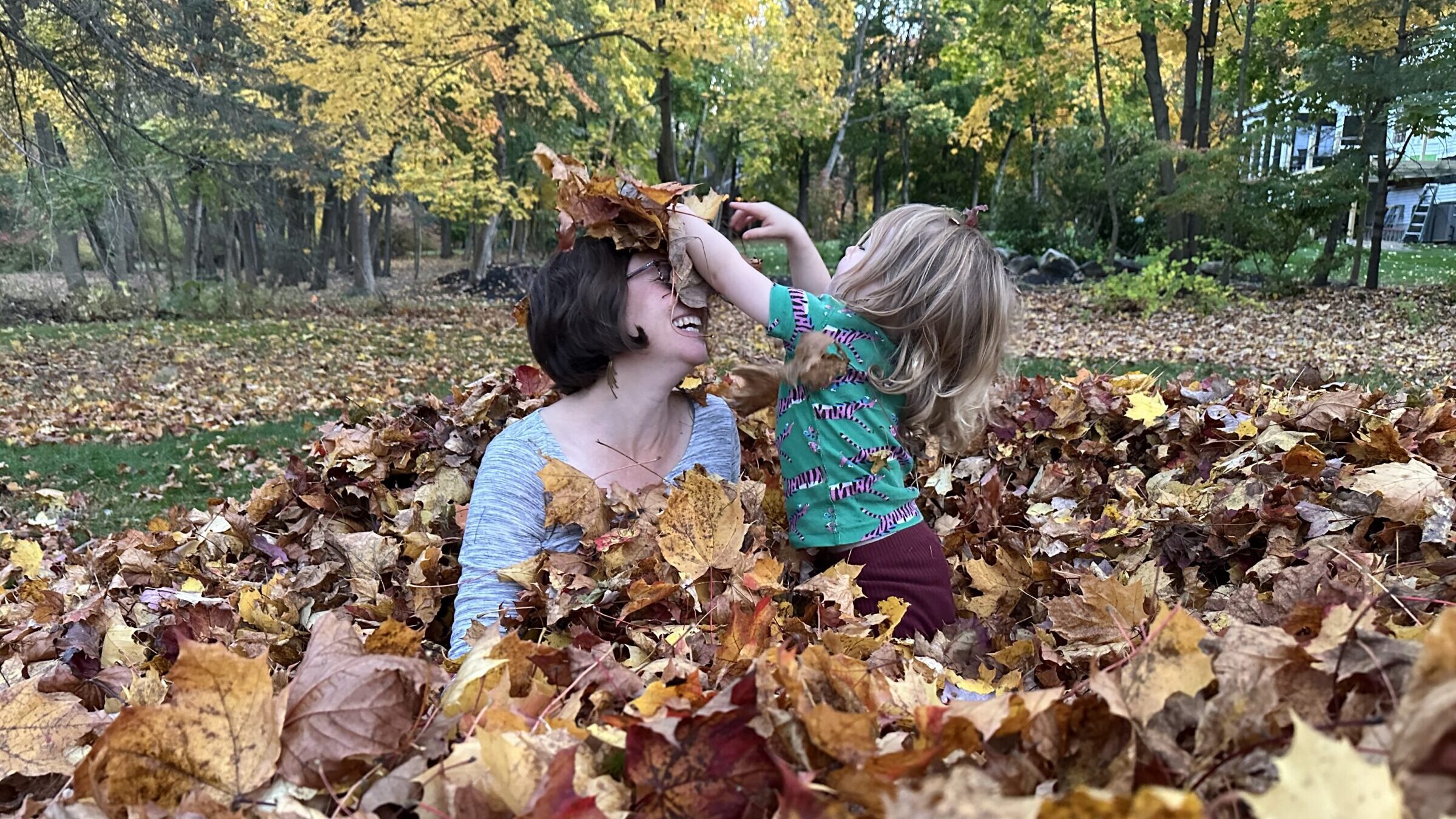 An adult and a young child laugh together while playing in a large pile of fallen maple leaves, surrounded by golden autumn trees.